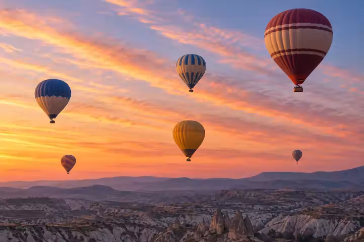 Six hot air balloons beneath coral sunrise clouds above Cappadocia’s valleys, with a burgundy and cream balloon in the foreground