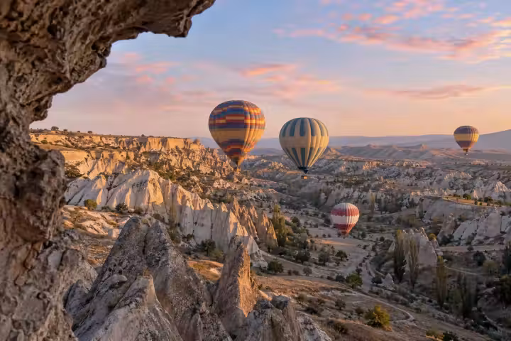 Four hot air balloons over a sunlit Cappadocia valley at sunrise, framed by a natural rock overhang