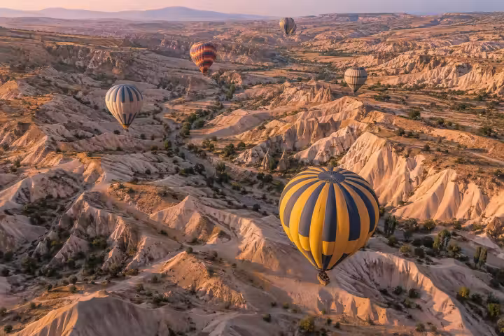 Hot air balloons above Cappadocia’s branching pale valleys at sunrise, seen from above with a yellow and blue balloon in the foreground