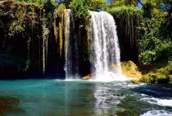 Broad white sheet of water falling past hanging moss and vines into a clear green pool, Antalya