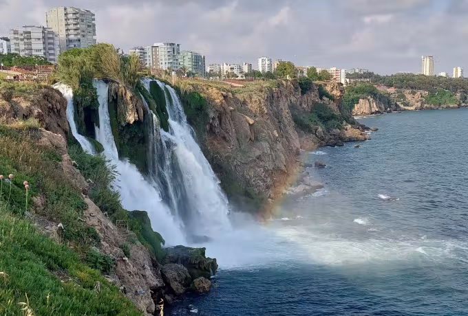 From the grass edge above Antalya: white streams down the rock face, a faint bow in the mist