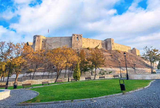 Gaziantep Kale on its mound over a cobbled walk and trees turned gold, bright cloud above