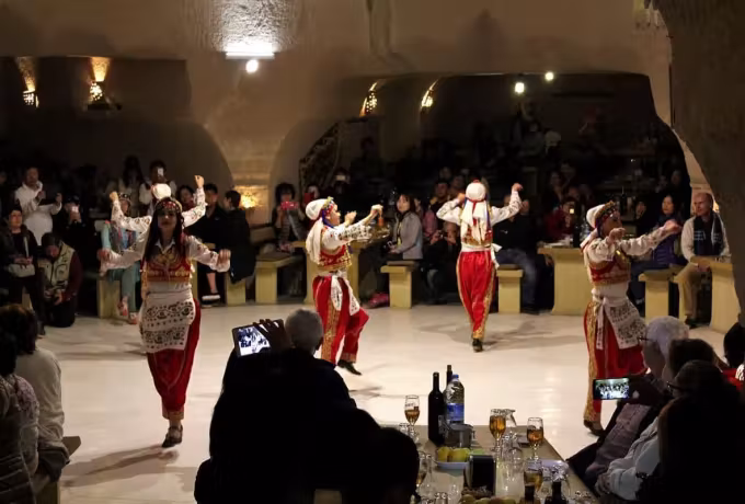 Troupe in ivory and gilt weaving past diners who lift cameras in a hewn Cappadocia room