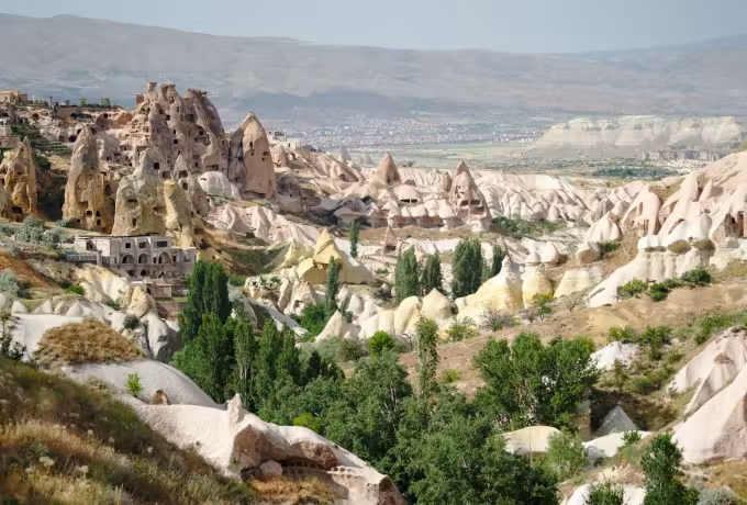 Broad view of hollowed dwellings and worn tufa with tall trees filling the bed under Uchisar