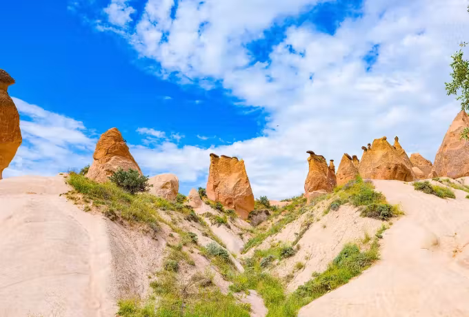 Columns in rose and flame tones rising out of scrub under bright sky with white cloud, Cappadocia