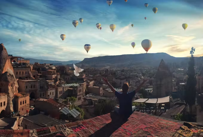Arms flung open on a rug-covered roof as envelopes drift over the cut-rock town below, Cappadocia