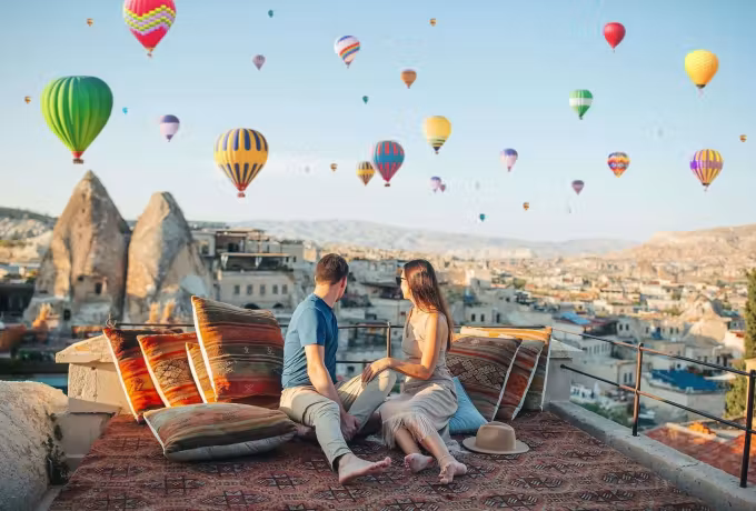 Pair on cushioned roof seating facing a crowded sky above the cut-rock houses, Cappadocia