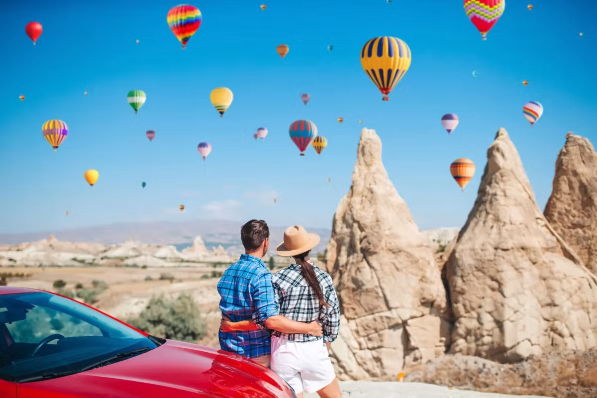 Pair leaning on a red car with heads tilted up at envelopes crossing above tall spires, Cappadocia
