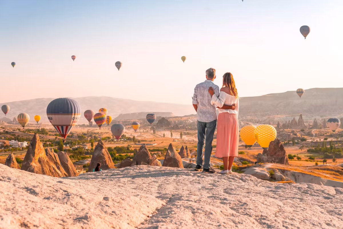 Standing together on bare stone above the spires as envelopes leave the valley in front, Cappadocia
