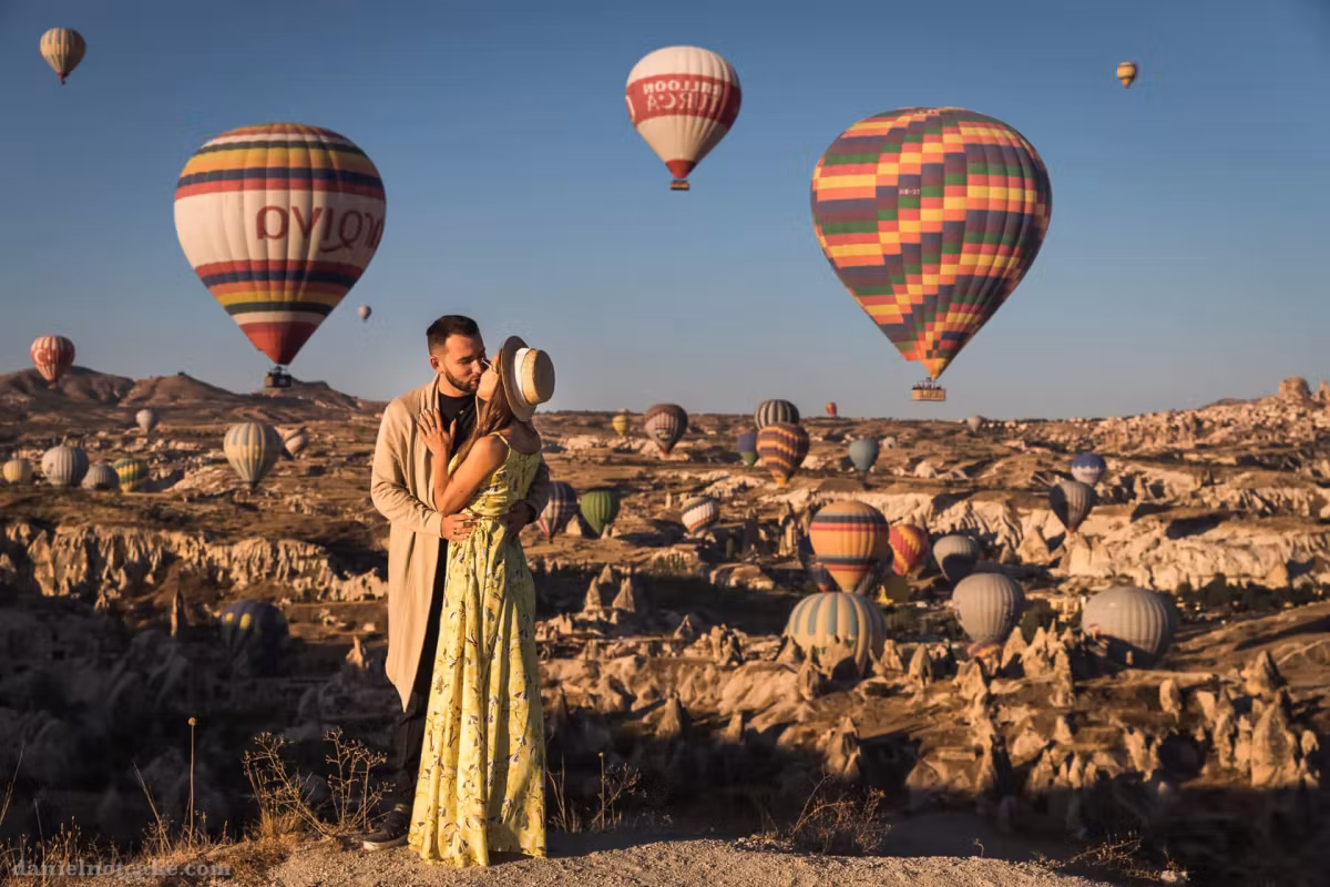 Couple holding each other with two big banded envelopes close behind and dozens far off, Cappadocia