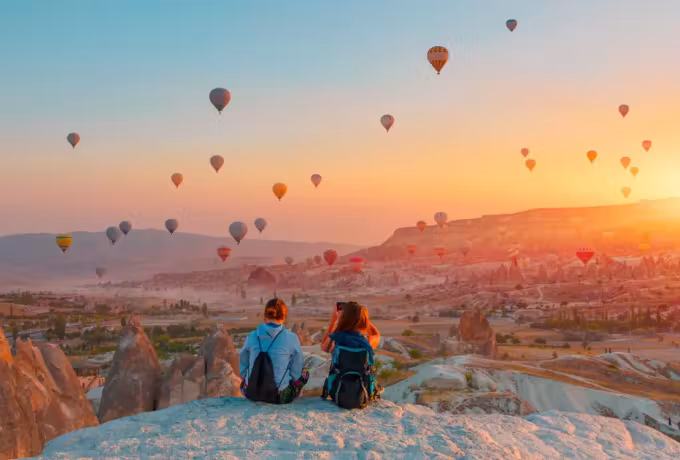 Two sat close on a pale stone crest as dozens of envelopes cross a blazing daybreak, Cappadocia