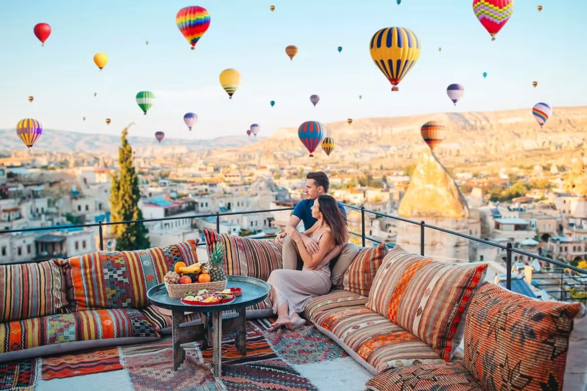 Pair at ease on a kilim bench below a sky thick with envelopes over the cut-rock town, Cappadocia