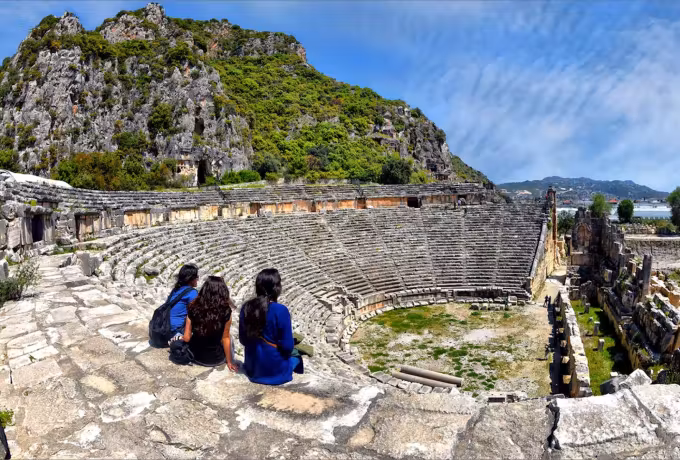Grey rock wall closing the back of the Roman auditorium at Myra, three people on the tiers