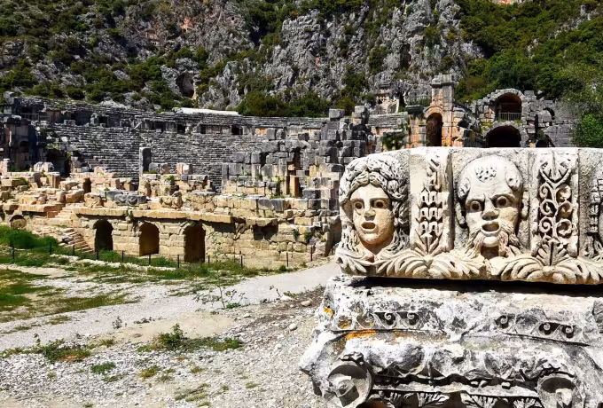 Fallen frieze at Myra carrying three deep-cut stage masks, arcaded walls and cliff beyond