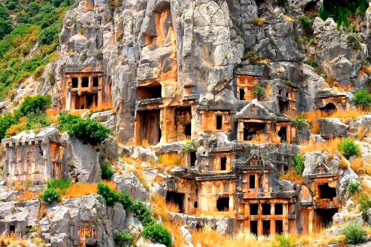 Tomb fronts with pedimented doors cut straight into the golden cliff at Myra, dry scrub below