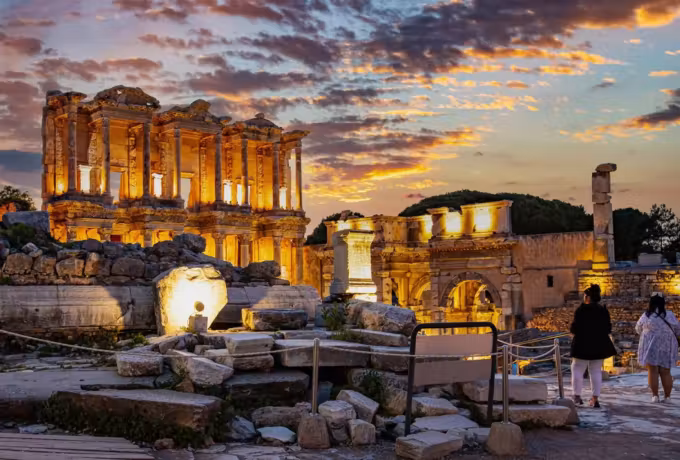 Evening burns orange behind the lit reading-room elevation, two onlookers down among the stones, Ephesus