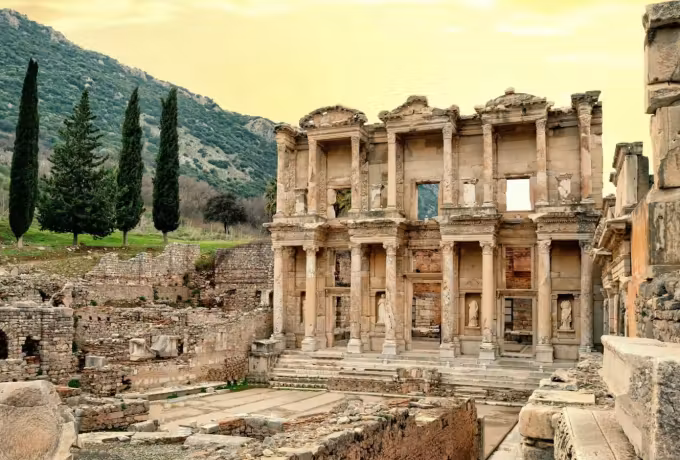 Masonry lit to a warm tone by the last sun, dark conifers climbing the bank alongside, Ephesus
