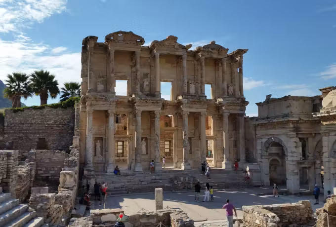 Soft noon light on the Celsus front, a palm rising over its left corner at Ephesus