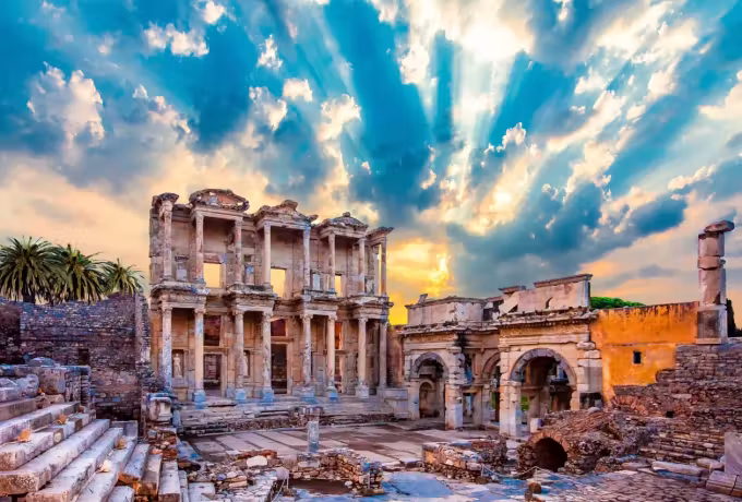 Shafts of light breaking heavy cloud and washing the Celsus front and gate warm orange, Ephesus