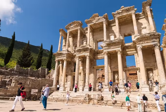 Brightly dressed people crossing the sunlit paving under the Celsus front on a clear day, Ephesus