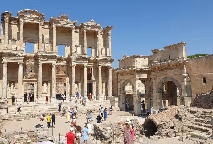 Unbroken traffic of sightseers passing the great elevation and arch under featureless noon light, Ephesus