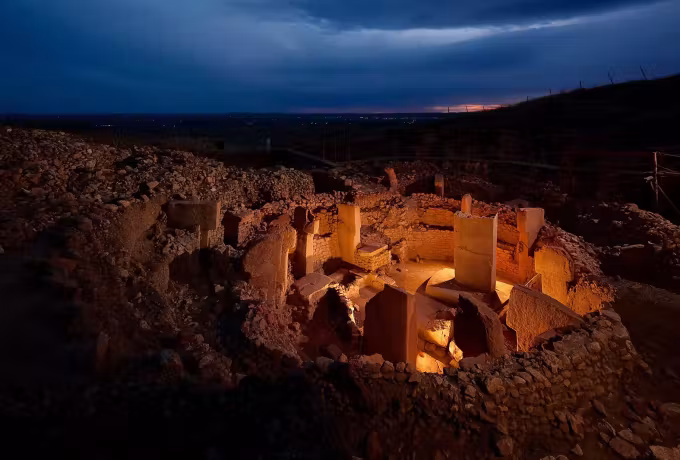 Evening lamps on the T-shaped uprights at Gobeklitepe, town lights faint across the plain