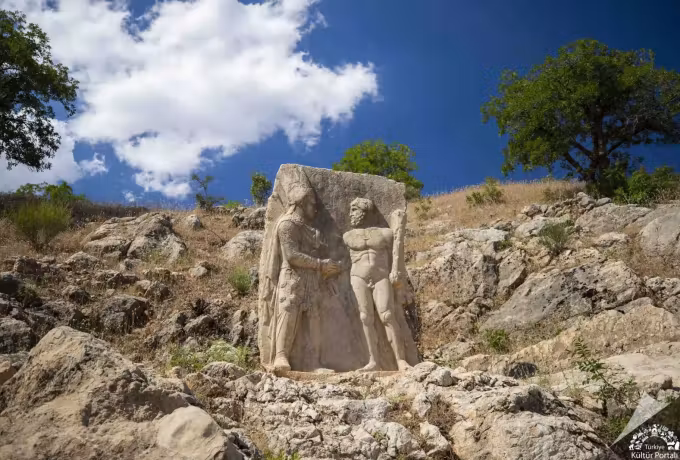 Robed ruler taking the hand of a club-bearing figure on a green hillside near Nemrut