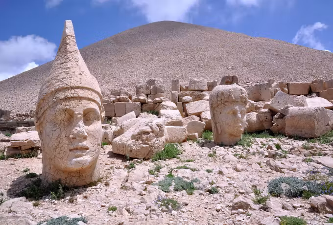 Head in a pointed cap under hard noon light, the smooth shingle mound at Nemrut behind