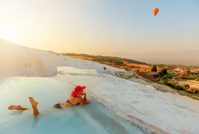 Red hat lying back in a shallow shelf pool while envelopes cross a pale morning sky, Pamukkale