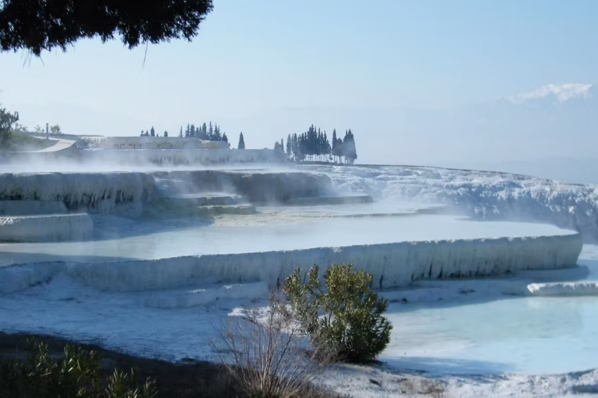 Steam lifting off warm shallows in freezing air, trees indistinct along the upper edge, Pamukkale