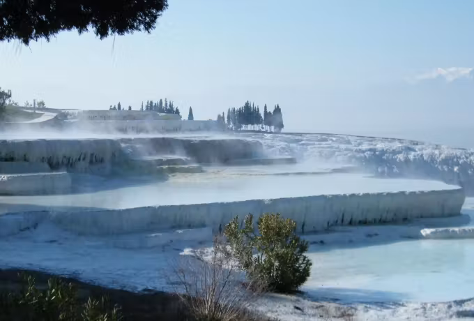 Steam lifting off warm shallows in freezing air, trees indistinct along the upper edge, Pamukkale
