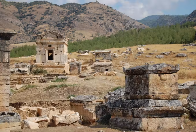Roofed sepulchre with a pediment in parched grass, timbered high ground beyond the cemetery, Pamukkale