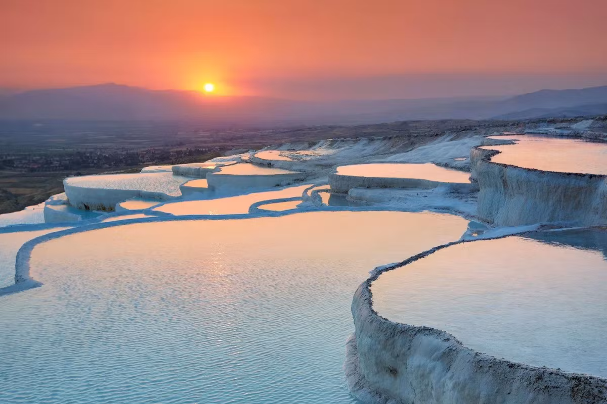 Sun sinking red over hazy country while the winding chalk lips glow warm below, Pamukkale