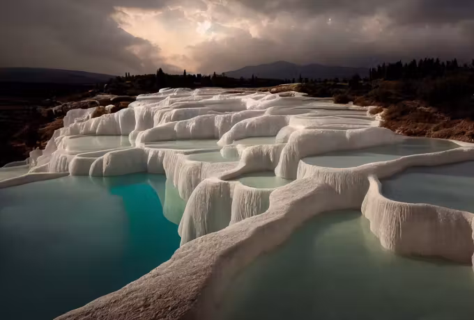 Rounded basins shining pale below heavy backlit cloud over the far hills, Pamukkale
