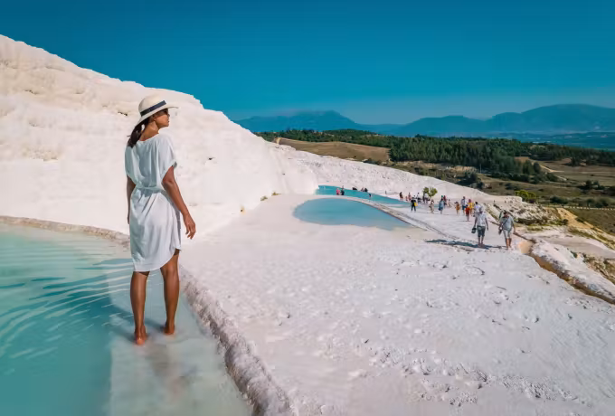 White dress and wide brim crossing wet chalk with people strung along the crest behind, Pamukkale