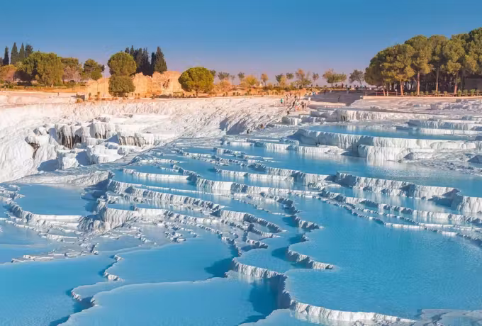 Thin chalk shelves holding blue water with pines and old walls along the plateau above