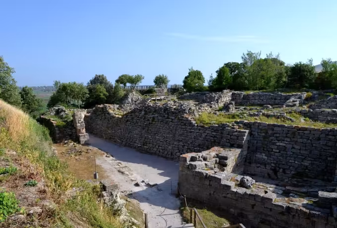 Dry-laid ramparts flanking a narrow cut passage at Troy, a visitor bridge crossing beyond