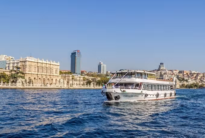 Ornate palace frontage on the strait with modern towers behind and a boat crossing deep blue