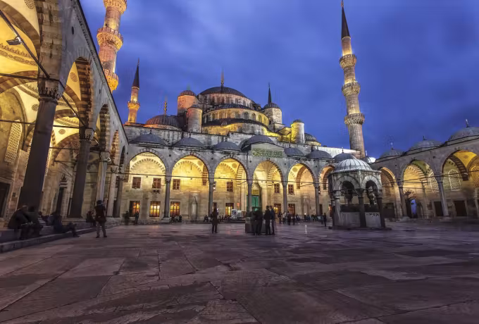 Lit forecourt viewed from under the arcading, opening toward cupolas and towers at dusk, Istanbul