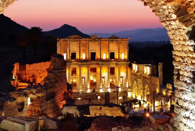 Celsus front glowing under night lighting, seen through a gap in a ruined wall at Ephesus