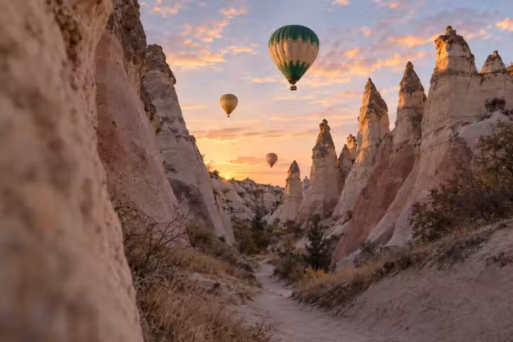Three hot air balloons above a winding path between tall Cappadocia rock formations in warm sunrise light