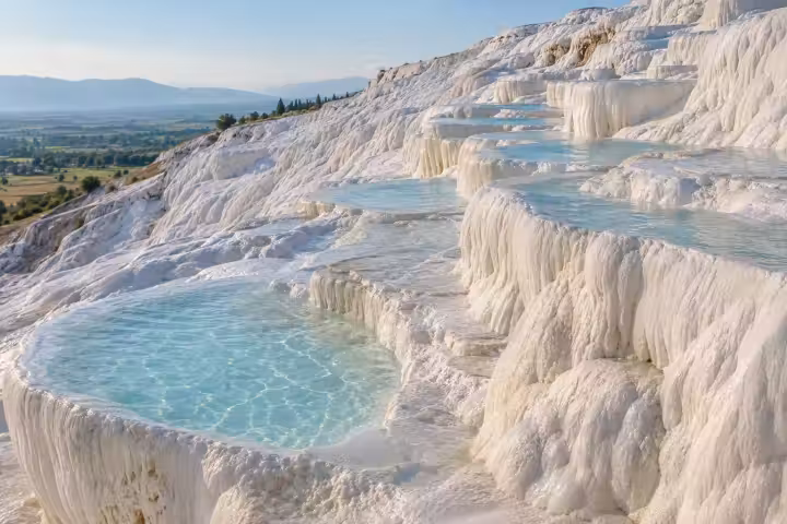 Scalloped white travertine terraces and pale blue pools at Pamukkale in morning light, with the green plain beyond