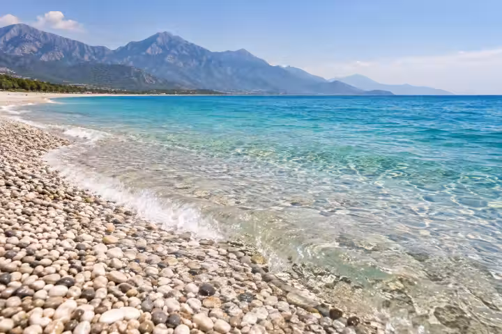 Clear turquoise water washing over a pebble beach on the Antalya coast, with mountains in the distance