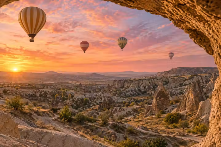 Four hot air balloons over Cappadocia at sunrise, framed by a natural rock opening