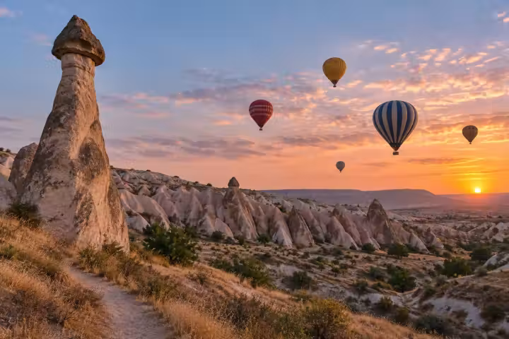 Five hot air balloons at sunrise beside a tall capped fairy chimney and valley trail in Cappadocia