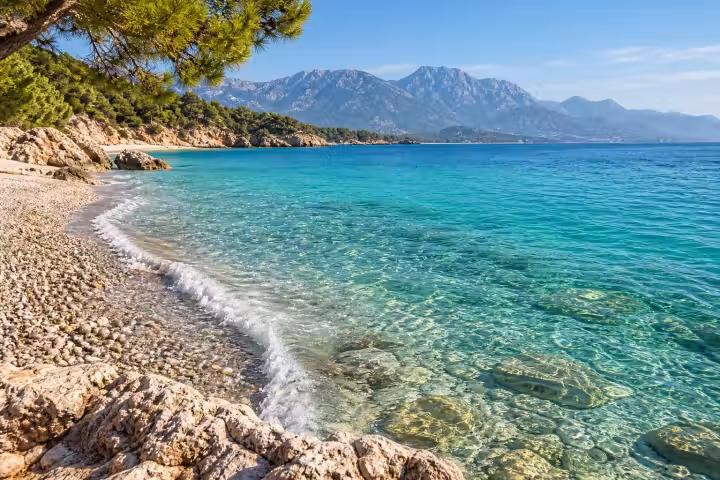 Clear turquoise water beside a pebble beach on the Antalya coast, framed by pine branches and mountains