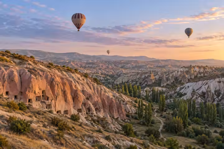 Hot air balloons over rose-coloured cave cliffs and a green Cappadocia valley at sunrise