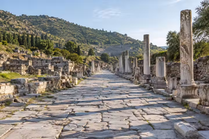 Weathered marble avenue and ancient columns beneath a green hillside at Ephesus