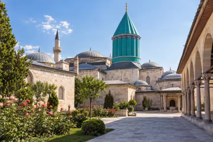Turquoise Mevlana Museum roof above a sunlit courtyard and rose garden in Konya